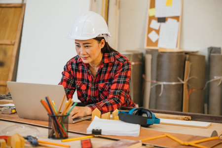 Carpenter woman in a white hardhat and red plaid shirt uses a laptop in a woodshop, surrounded by tools, wood scraps, and pencils. Ideal for online learning, client orders, or woodworking projectsの写真素材