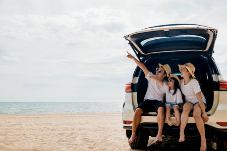 Family Day. Father, Mother and daughter enjoying road trip sitting on back car and point finger out blue sky, Happy people having fun in summer vacation on beach, Family travel in holiday at sea beachの写真素材