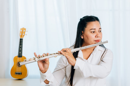 A female flutist in a white shirt sits on a chair in her living room, playing a silver flute. The soft curtain backdrop enhances the serene atmosphere as she passionately plays the musical instrument.の写真素材
