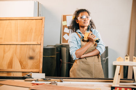 Smiling carpenter woman holding a power drill in a workshop, with a wooden stool and small house models nearby. Surrounded by tools, showcasing her craft and enthusiasm for woodworking projectsの写真素材