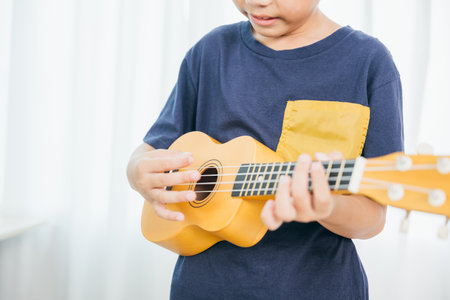 A detailed shot of a young boy strumming a yellow ukulele in a cozy living room. His blue shirt contrasts with the soft white curtain backdrop, emphasizing his focus on the music he loves to play.の写真素材