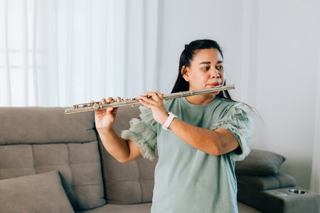 A female flutist in a green shirt sits gracefully on gray sofa, playing a silver flute. The cozy living room setting adds warmth and serenity, showcasing her passion for music in a casual environment.の写真素材