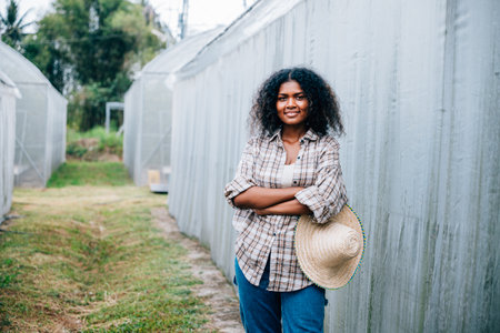 Confident farmer woman in checkered shirt and apron arms crossed smiling at the camera amidst vibrant greenhouse tomatoes. A portrait reflecting happiness and care in farm horticulture.の写真素材