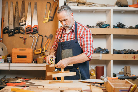 Senior carpenter uses tape measure to measure wooden slats for project, ensuring precision in each cut. Craftsman focuses on DIY woodworking and home renovation, with tools and materials in backgroundの写真素材