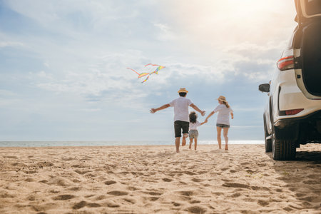 Asian family is flying a kite while running on the beach. Happy parents mother and father with their child playing with kite, The car is parked nearby, and the family is enjoying their time togetherの写真素材