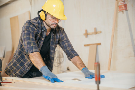 Young carpenter in a yellow helmet, blue gloves, and plaid shirt using a circular saw to cut wood in a workshop. Ideal for showcasing woodworking skills and construction techniques.の写真素材