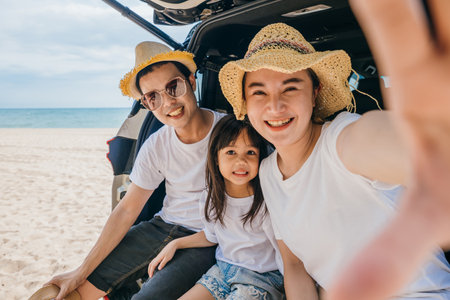 Happy Asian family enjoying having fun while taking a selfie with smartphone while sitting on back end of a white SUV parked on a beach background, Summer trip road conceptの写真素材