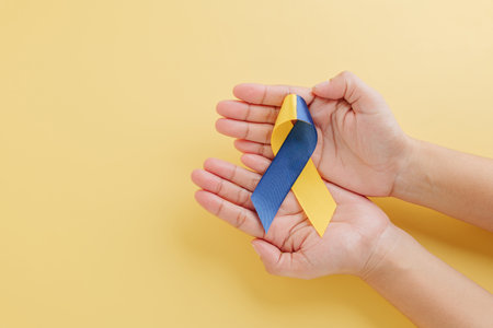 Two hands gently hold a yellow and blue awareness ribbon on yellow background, symbolizing Down Syndrome Day. Promoting awareness for Down Syndrome and Autism Awareness globally.の写真素材