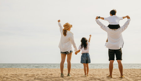 A joyful family of four stands with raised arms at the sea beach during sunset. Father carries his...