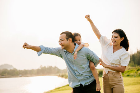 Asian family child girl piggyback riding back on father raising hands like flying outdoor nature park with mother, Happy parent daughter having fun together, Tourist lifestyle family leisure activityの写真素材