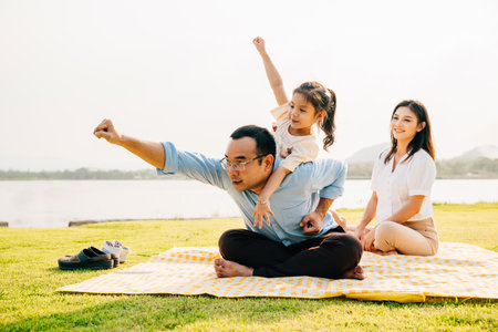 Asian family sitting playing on picnic blanket spring meadow field grass in park, Happy parents child girl riding father back enjoying outdoors together, father carrying daughter, Family relaxationの写真素材