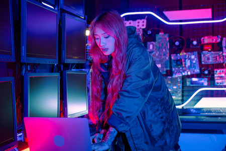 Asian woman in front of a computer monitor display with laptop computer, neon light, wear black jacket at system control room, working on multiple monitorsの写真素材