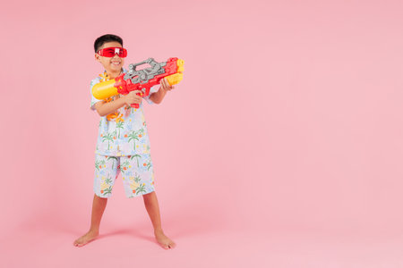 Songkran festival fun full body cheerful child in a floral shirt and glasses holds a water gun, celebrating the traditions and excitement of Songkran in Thailand, happy smile isolated pink backgroundの写真素材