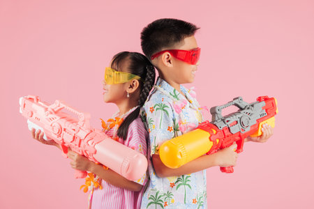Songkran festival fun as two Thai kids, a boy and a girl in glasses, hold water guns together, capturing the playful spirit of Songkran in Thailand with bright smiles, isolated pink backgroundの写真素材