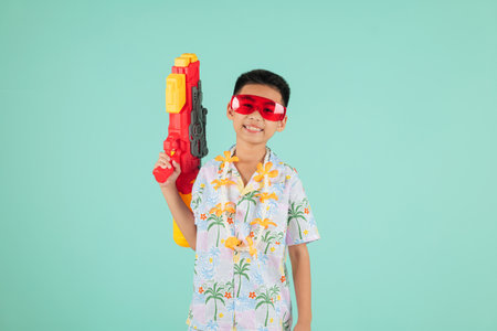 Songkran festival thrill as a happy boy in glasses and a floral outfit holds a water gun, enjoying the excitement of Songkran in Thailand with cultural traditions, isolated blue backgroundの写真素材