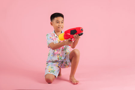 Songkran happiness full body as a cheerful Thai child sits holding a toy water gun, dressed in a floral outfit, enjoying the fun and culture of Songkran celebrations, isolated pink backgroundの写真素材