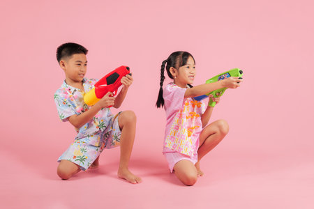 Songkran excitement shines as a full body Thai boy and girl sit together, holding plastic water pistols, enjoying the festive traditions of Songkran in Thailand, isolated pink backgroundの写真素材
