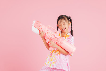 Songkran joy with a happy girl wearing a floral shirt holding a toy water gun, enjoying the playful Songkran festival atmosphere in Thailand with a bright and cute smile. isolated pink backgroundの写真素材