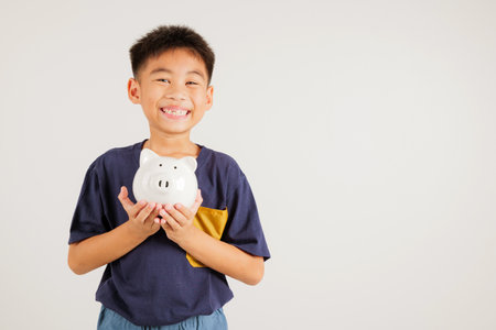 Asian little kid boy holding piggy bank and looking at camera at studio shot isolated on white background, Happy primary child lifestyle smiling with piggybank, Thai personal money savingsの写真素材