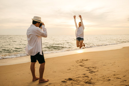 Love in focus as young couple dressed in white enjoys sunrise date on beach. boyfriend armed with camera lovingly captures snapshots of his girlfriend creating memories by ocean.の写真素材