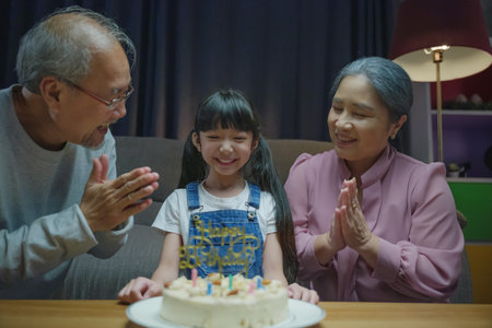 Happy Asian young girl celebrating her birthday by blowing out candles on a cake with grandparent, elderly grandma and grandpa congratulating granddaughterの写真素材