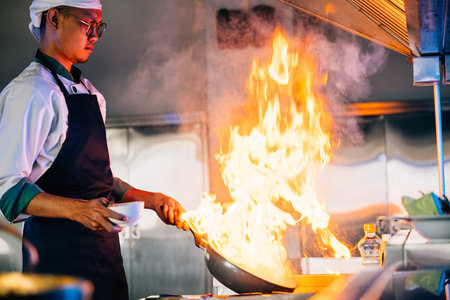 Chef hands with wok and fire. Closeup of chef cooking food with flames in professional kitchen. Expertise at work busy cooking handling flames modern kitchen environment. cook food with fireの写真素材