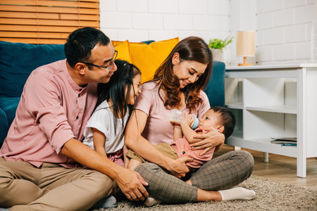 In the comfort of their home a mother feeds her newborn baby with a bottle while her toddler son and daughter join in bringing happiness and togetherness to their family portrait.の写真素材