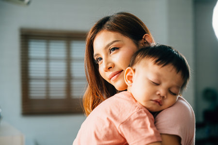 In the peaceful setting of their home an affectionate Asian mom leans in to embrace and cuddle her small sleeping infant celebrating a heartwarming family moment filled with the joys of parenthood.の写真素材