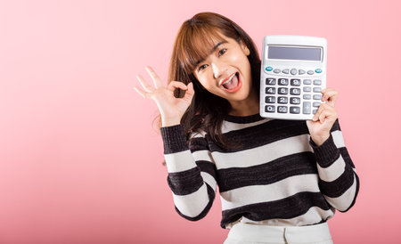 Tax day concept. Woman confident smiling holding electronic calculator and show OK gesture, excited happy Asian female studio shot isolated on pink background, Account and finance counting incomeの写真素材