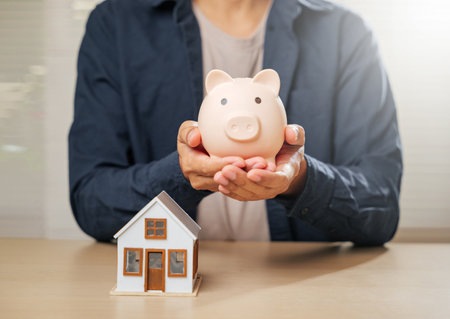 A close-up of hands with a piggy bank and house model, symbolizing saving money, financial planning, and property goals. Highlights mortgage options and homeownership strategies for a secure future.の写真素材