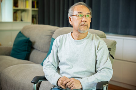 Asian senior man disabled sitting alone on wheelchair in living room looking out window at home and thinking, lonely elder sad old man retirement memory loss or nostalgia thought, feel depressedの写真素材