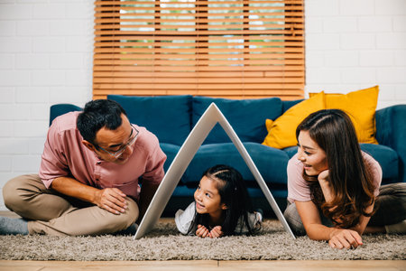 In their new home a cheerful family on a couch holds a cardboard roof symbolizing happiness. Asian parents and their daughter showcase security and togetherness.の写真素材