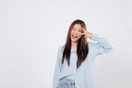 Victory gesture near eye with smile, showing peace and joy. Victory mood captured isolated on white background, optimistic Asian woman expresses positive vibes and excitement.の写真素材