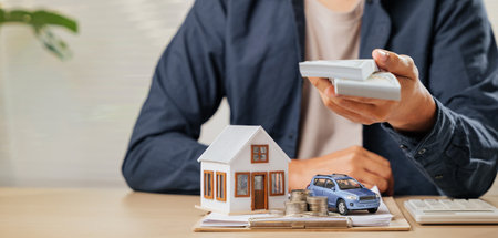 Money bills held above house and car models with paperwork. Represents tax-saving strategies, cash flow, and property loan planning for financial stability and asset acquisition.の写真素材
