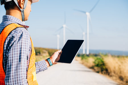 Man in hardhat using tablet at windmills. Engineer assures clean energy generation. Expertise in...