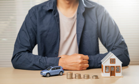 Man sitting with small car and house models beside coin stacks on a table. Concept of finance, saving for property, auto loans, down payments, installment costs, and tax strategies for homeownership.の写真素材