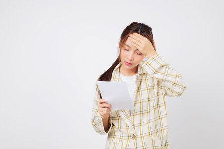 Financial stress overwhelms the Asian woman as she holds a bill in each hand, reviewing her financial troubles and unpaid bill stack with a worried face. isolated studio white backgroundの写真素材