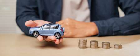 Hand holding a blue toy car model next to ascending coin stacks. Depicts auto credit, car loans, cash installment payments, and financial planning for managing car purchase costs.の写真素材