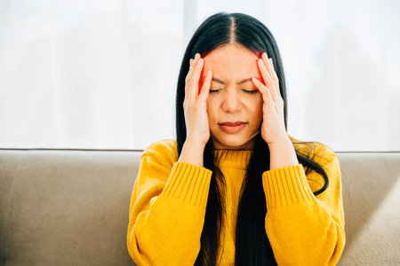 Seated on a sofa an Asian woman manifests stress and headache touching her face. Illustrating health care and medical concept revealing discomfort and exhaustion.の写真素材