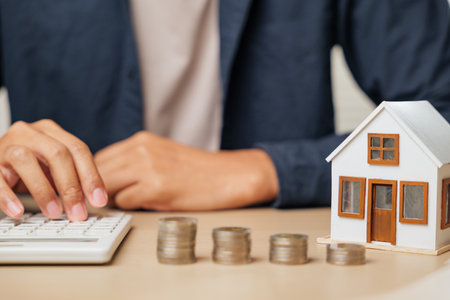 Person using calculator next to house model and cash stacks. Reflects real estate and property tax planning, profit increase strategies, and financial decisions for people aiming for smart investmentsの写真素材