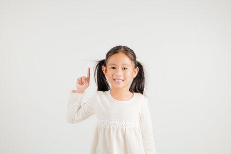 Asian happy portrait cute young kid girl makes gesture fingers point upwards above presenting product something, studio shot isolated on white background, kindergarten children woman smileの写真素材