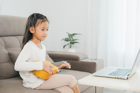 A young girl practicing ukulele on a couch, using a laptop for online tutorials. This scene illustrates the convenience and innovation of music education in a personalized, home setting.の写真素材