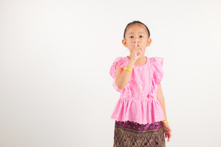 Portrait Thailand kid girl smiling traditional Thai dress costume making finger on lips mouth silent quiet gesture, studio isolated white background, kindergarten child marking silence for stop quietの写真素材