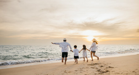A happy Asian family seen from behind enjoys sunny day at beach. Parents and daughters bond running jumping and pretending to fly with arms outstretched. carefree family moment by sea.の写真素材