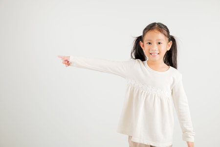 Happy Asian portrait cute young kid girl smiling indicate finger empty space, studio shot isolated on white background, Thai kindergarten child pointing index out with copy spaceの写真素材