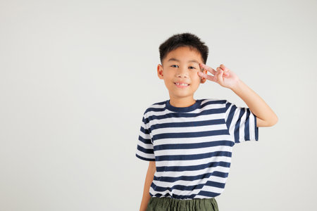 Asian young kid boy showcases optimism and peace by gesturing victory and peace signs in a studio shot with a white background. primary child joyful smile speaks of success, V sign fingerの写真素材