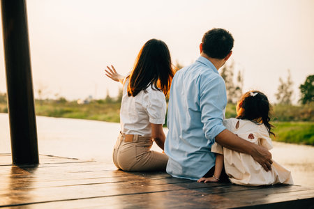Asian family of three a girl child are sitting on a wooden dock by a body of water. Happy parent father mother and daughter kid they are all smiling. Concept of family warmth and happinessの写真素材