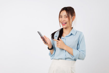 Asian woman holding phone in one hand while pointing happily at the phone screen, feeling excited and smiling with joy in a casual moment full of fun and expression. isolated studio white backgroundの写真素材