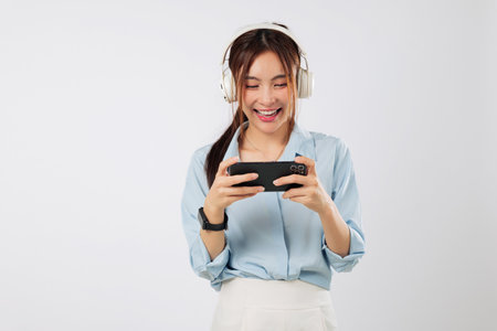 Game vibes and game joy shown by excited Asian woman playing on mobile phone with headphones, smiling and enjoying funny online video. isolated studio white backgroundの写真素材