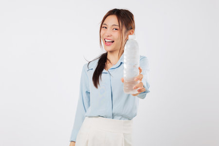 A young asian woman holds a plastic bottle with water, showing a smile as she prepares to drink the water in her hand for healthy hydration and refreshment, isolated studio white backgroundの写真素材